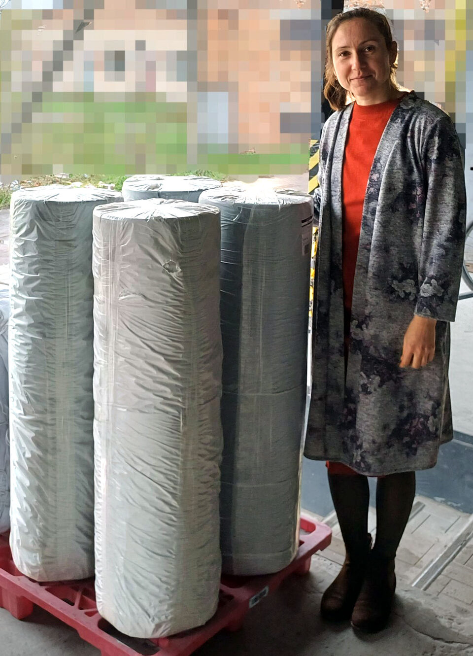 A volunteer stands beside four tall rolls of fabric wrapped in plastic on a red pallet